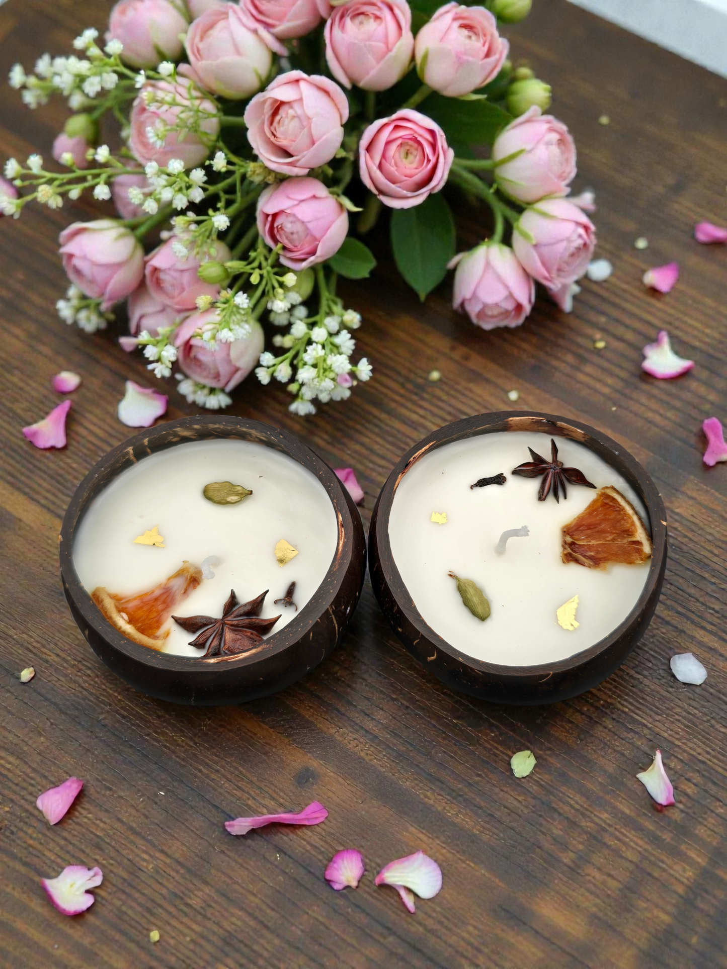 Two small candles with decorative elements on a wooden surface with pink flowers and petals.