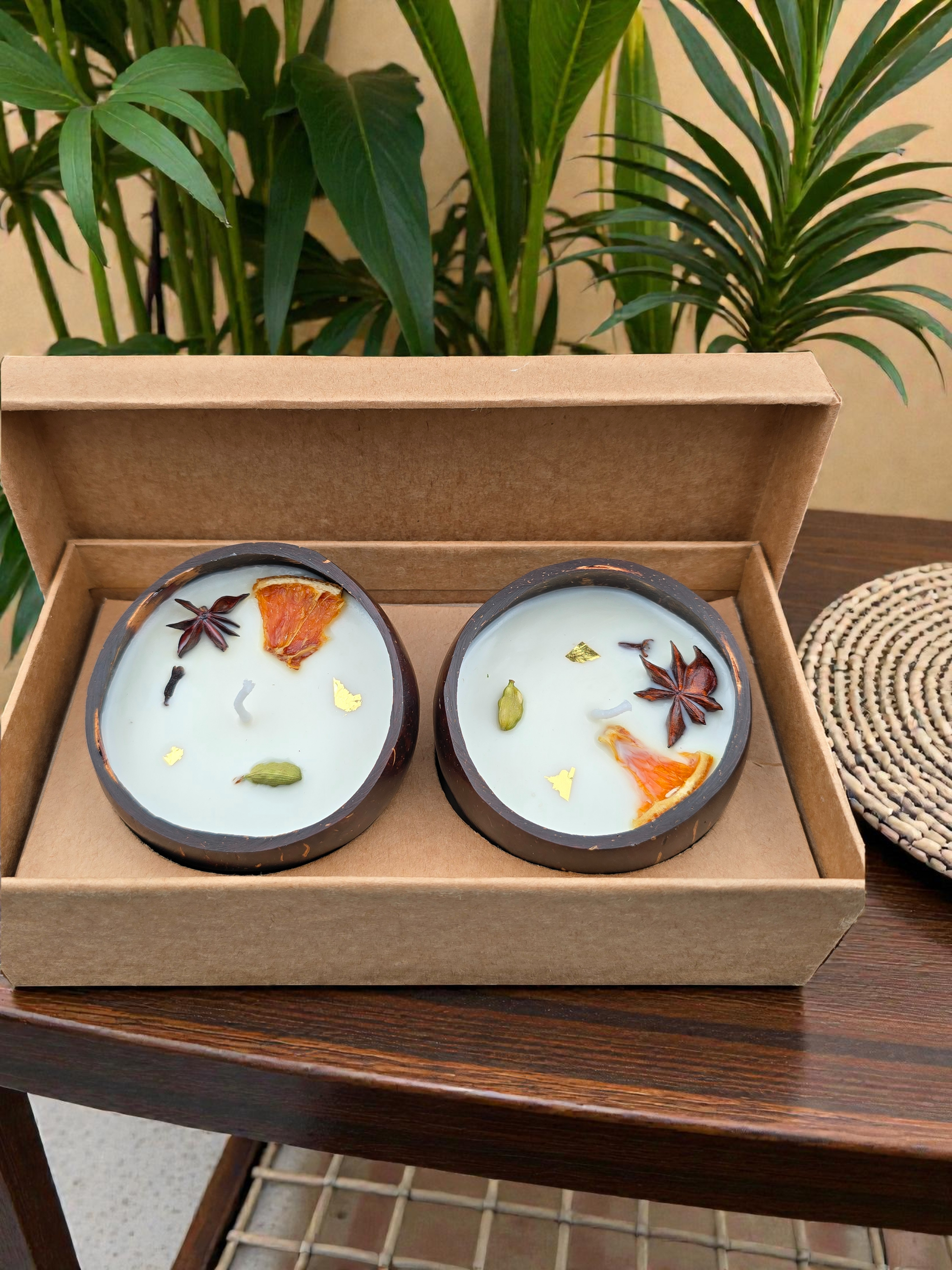 Two candles with decorative elements in a cardboard box on a wooden table with plants in the background.