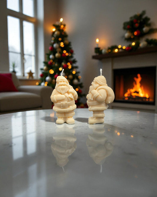 Two Santa Claus-shaped candles on a reflective surface with a festive background including a Christmas tree and fireplace.